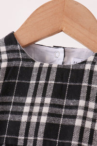 A close-up of a black and white plaid garment hanging on a light wood hanger against a plain light background.