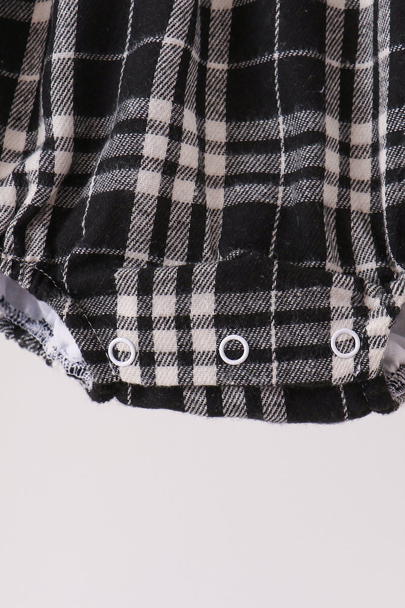 Close-up of a black and white plaid garment, likely a baby romper or shorts, showing three white snap closures along the bottom hem against a light gray background.