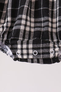 Close-up of a black and white plaid garment, likely a baby romper or shorts, showing three white snap closures along the bottom hem against a light gray background.