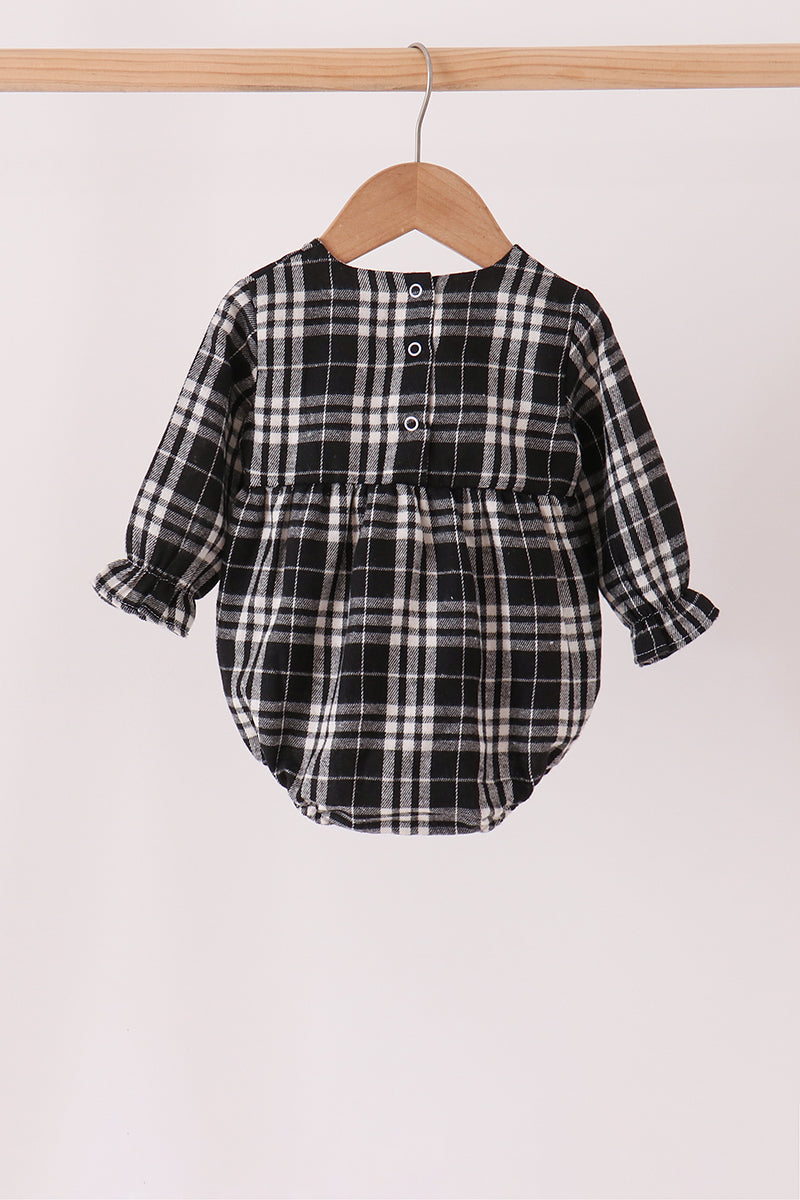 A baby long-sleeve romper in a black, white, and gray plaid pattern, hanging on a wooden hanger from a wooden clothes rack against a plain off-white background.