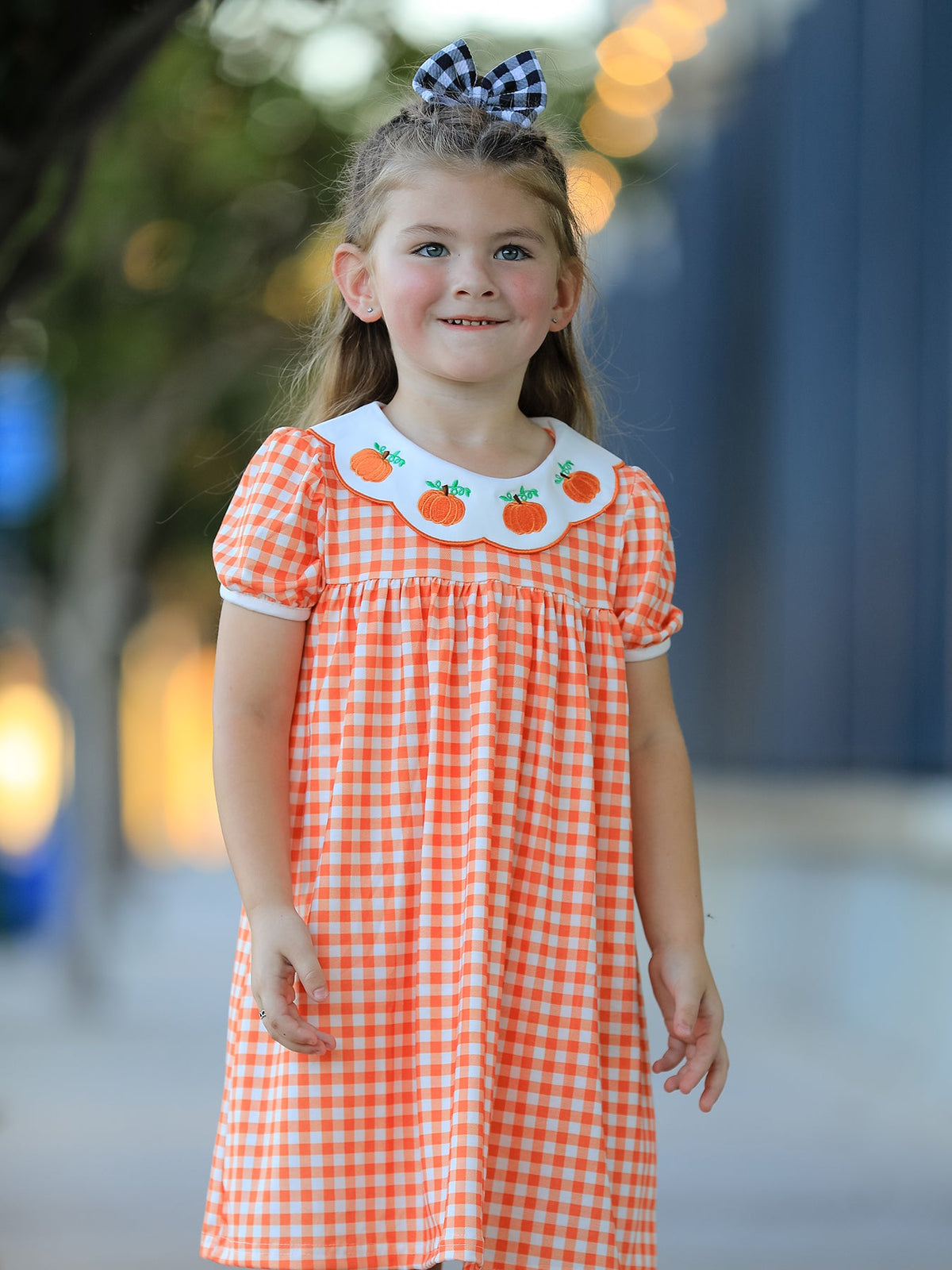 Girl wearing orange gingham puff sleeve dress with pumpkin embroidered collar and black white buffalo check bow.