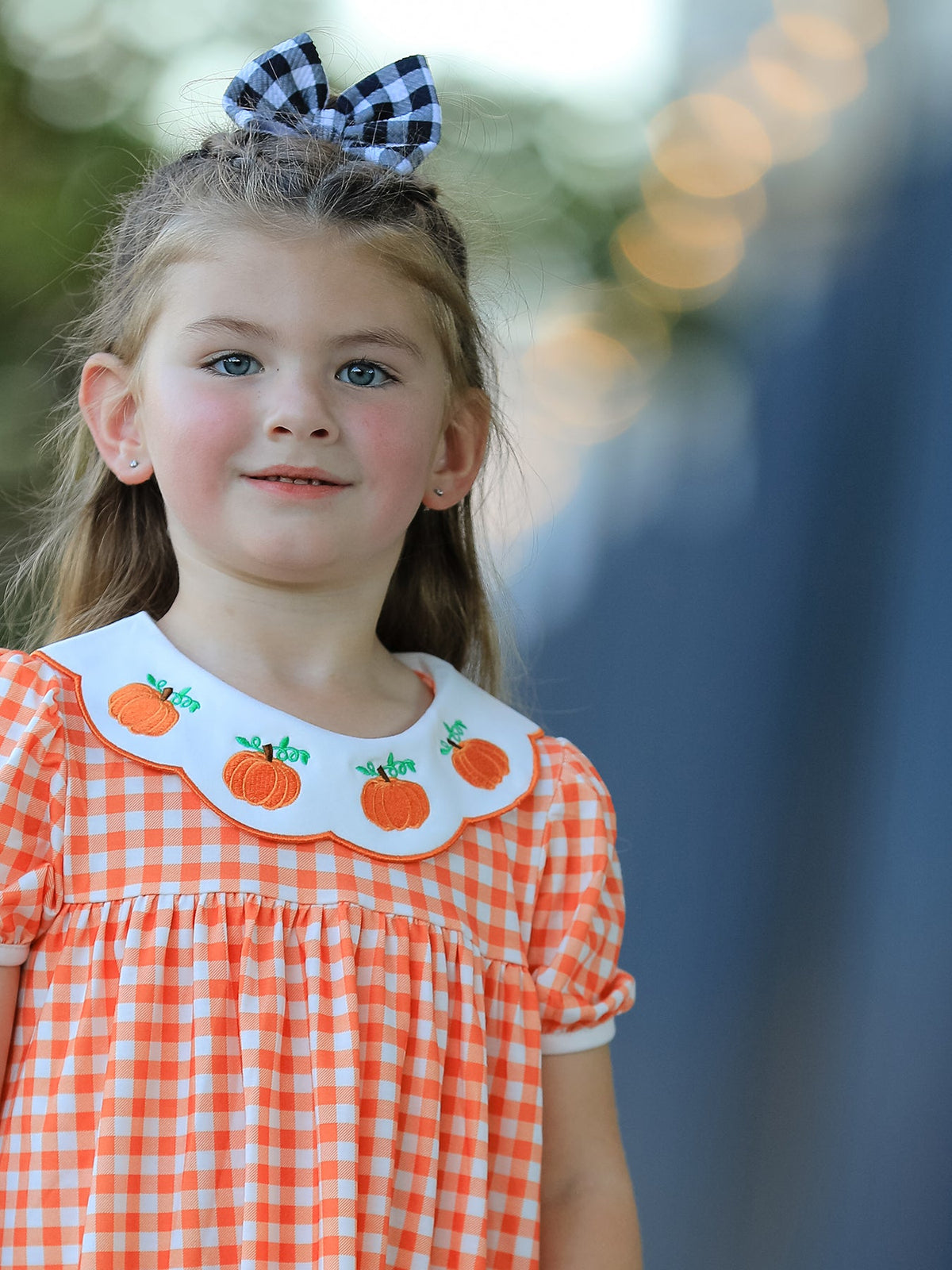 Orange gingham puff sleeve dress with pumpkin-embroidered white collar worn by a smiling girl with a buffalo plaid bow.