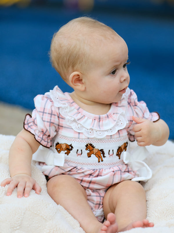 Baby girl wearing pink plaid romper with embroidered horses and smocked bodice. Western cowgirl style one-piece with ruffled lace collar.