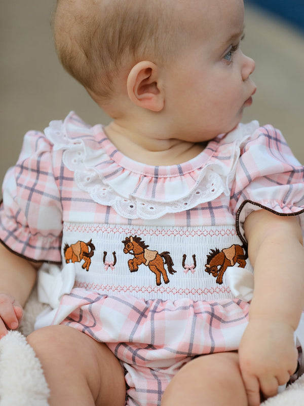 Baby girl wearing pink plaid romper with embroidered western horses and horseshoes on a smocked white bodice and lace collar.