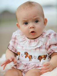 Baby girl wearing pink plaid romper with embroidered Western horses and smocked bodice. Features ruffled lace collar and ranch-style design.