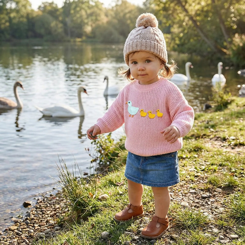 Toddler wearing pink embroidered duck sweater, beige beanie, denim skirt by lake with swans