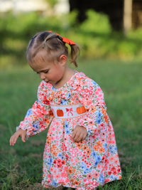 Young girl with orange hair bow wearing a fall floral Pumpkin Bow Smock Dress with embroidered pumpkins, outdoors.