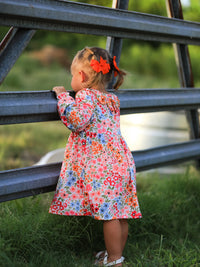 Little girl in floral Fall Pumpkin Bow Smock Dress with orange hair bows, standing by a metal railing.