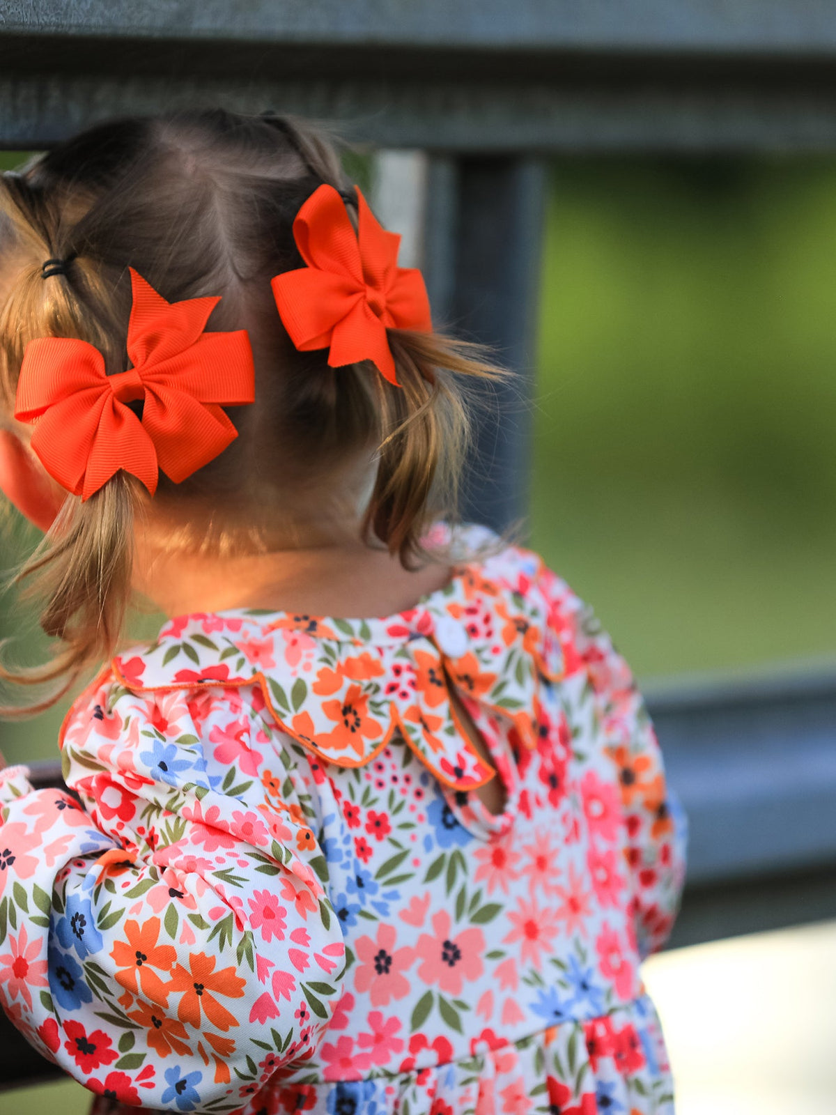 Girl seen from behind with two bright orange pumpkin bows in hair, wearing a floral smock dress.