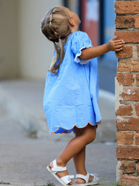 Blue fall dress for baby girl with dotted fabric, lace sleeves and orange scalloped hem beside a brick wall.