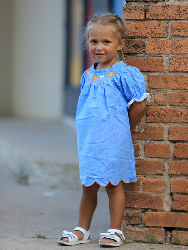 Blue smock baby girl dress with pumpkin embroidery and lace trim, worn by a toddler beside a brick wall.