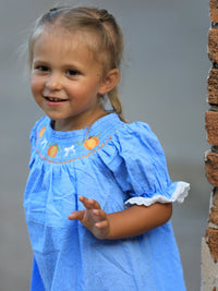 Smiling toddler girl in blue fall dress with pumpkin embroidery, lace sleeves, and smocked bodice.