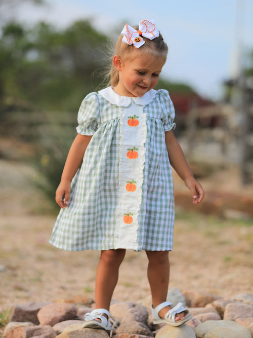 Young girl in green plaid dress with pumpkin appliques and a pumpkin bow, outdoors in fall.