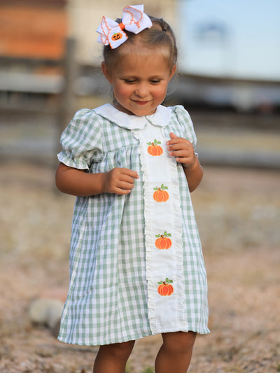 Young girl in green plaid dress with pumpkin appliques on white placket, outdoors.