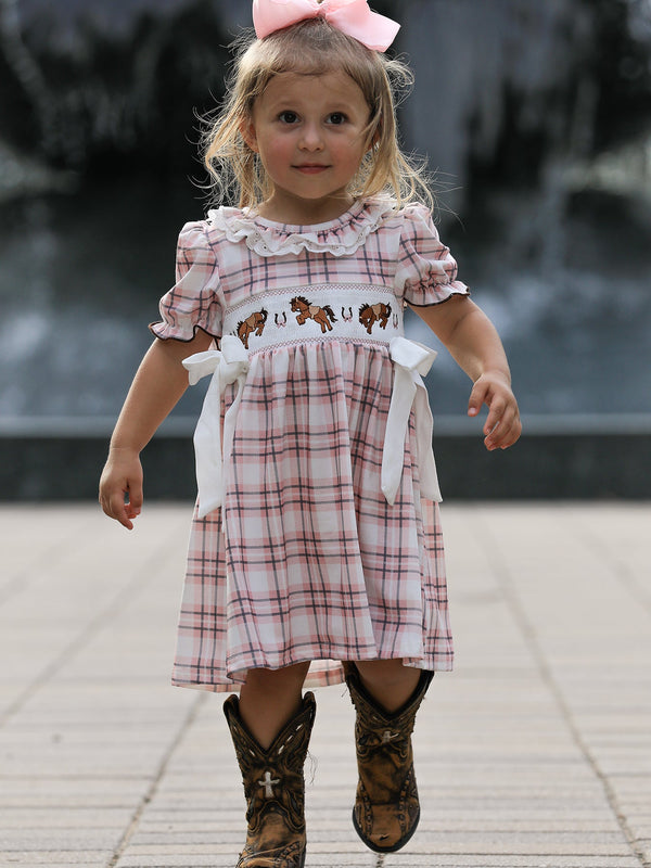 Toddler girl wearing pink plaid Western dress with embroidered horses, white bows, and cowboy boots.