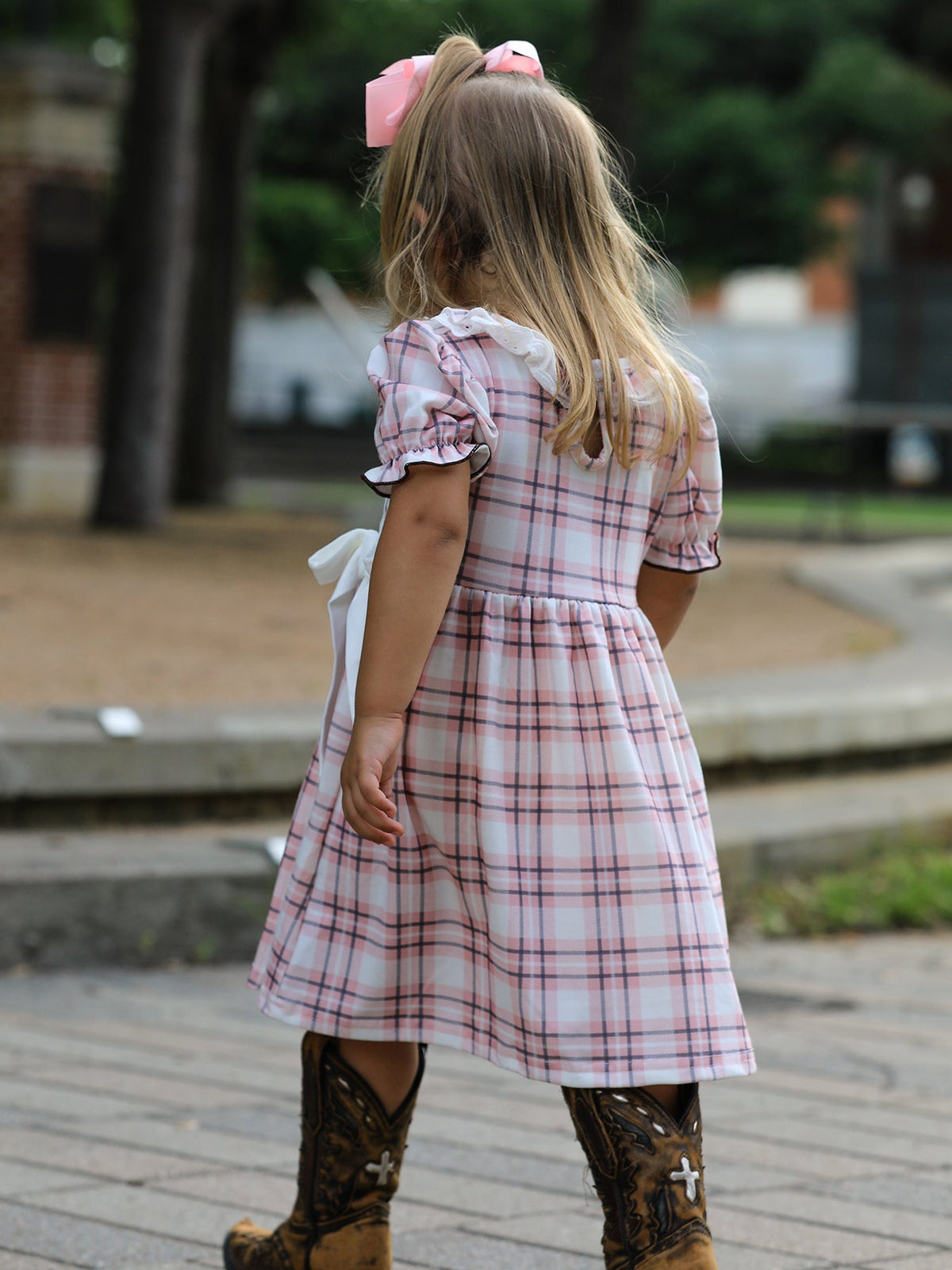 Back view of a girl wearing a pink and white plaid Western dress with ruffled sleeves and brown cowboy boots.