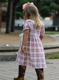 Back view of a girl wearing a pink and white plaid Western dress with ruffled sleeves and brown cowboy boots.