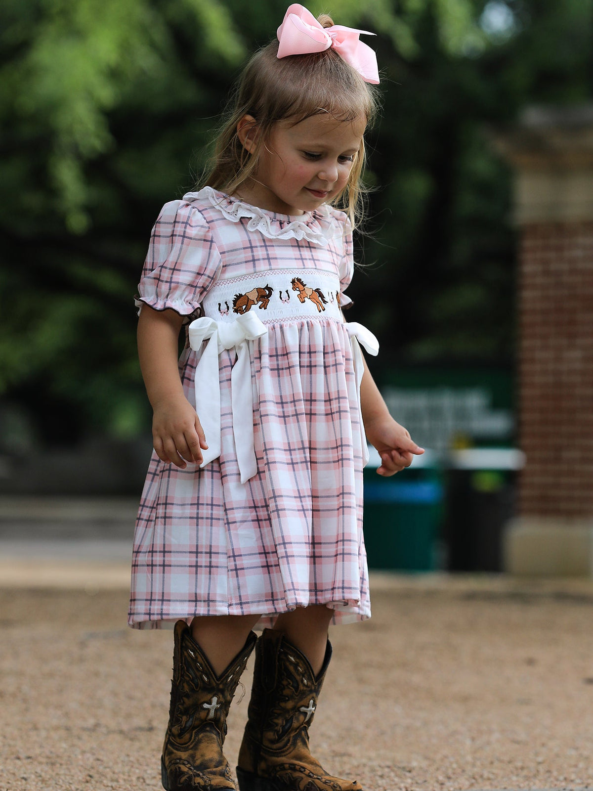 Toddler girl in pink plaid Western dress with horse embroidery, smocked bodice, white bow, and cowboy boots.