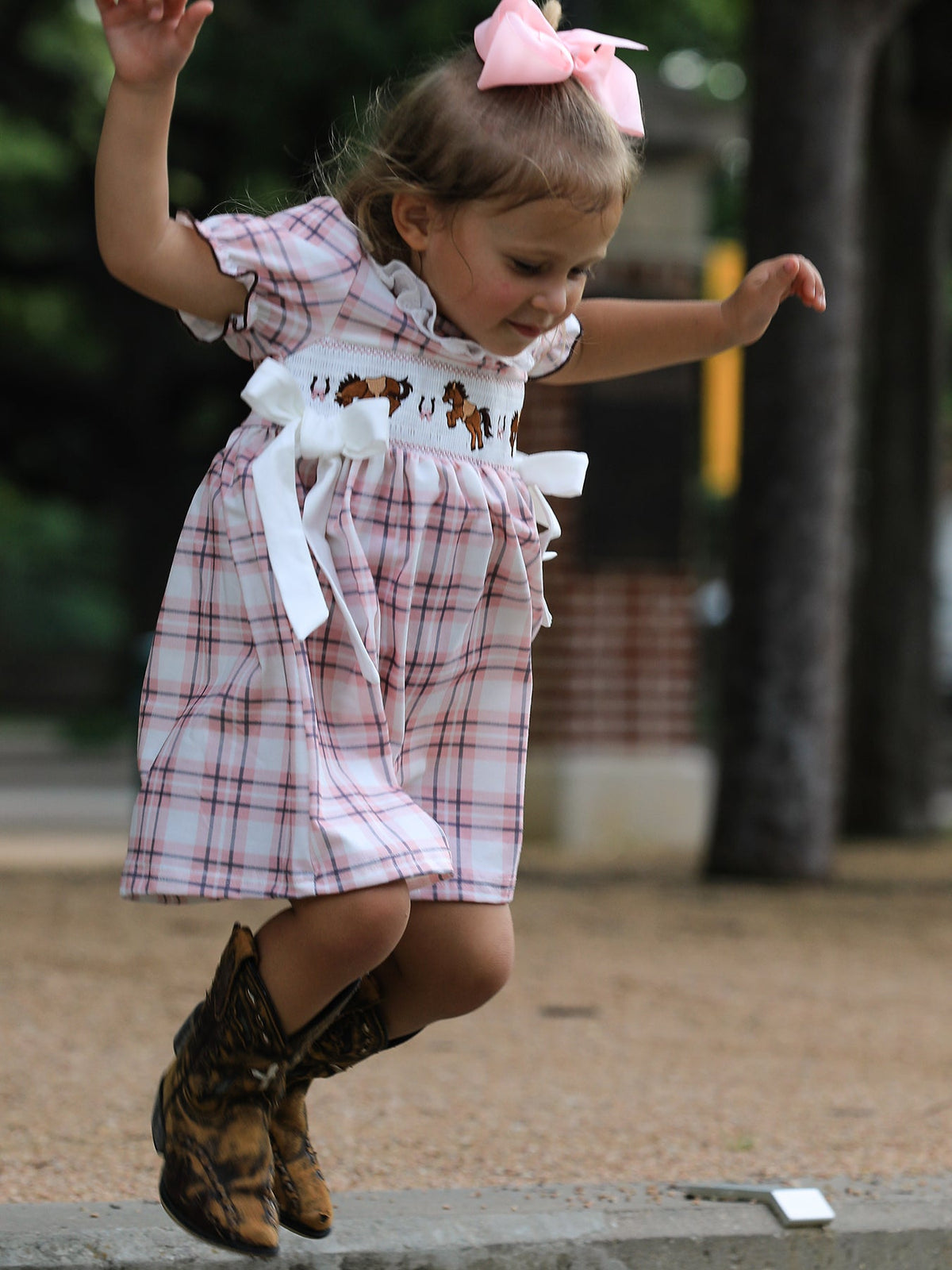 Toddler girl jumps in pink plaid Western dress with horse embroidery, smocked bodice, white bows, and cowboy boots.