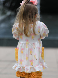 Back view of a girl in a white dress with pink bow prints and yellow side pockets, paired with orange ruffled shorts.