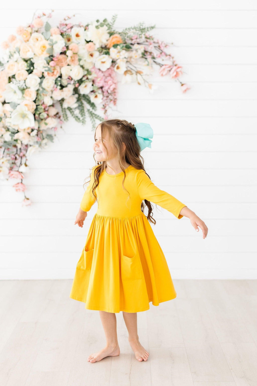 A joyful girl wearing the Mustard 3/4 Sleeve Pocket Twirl Dress by Mila & Rose®, twirling with happiness. The dress features a playful twirl skirt and convenient pockets. Set against a backdrop of beautiful florals, she radiates fun and adventure.