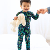 Child wearing Mystic Mama blue Second-Skin Convertible Romper, holding a teddy, with caregiver's hand.