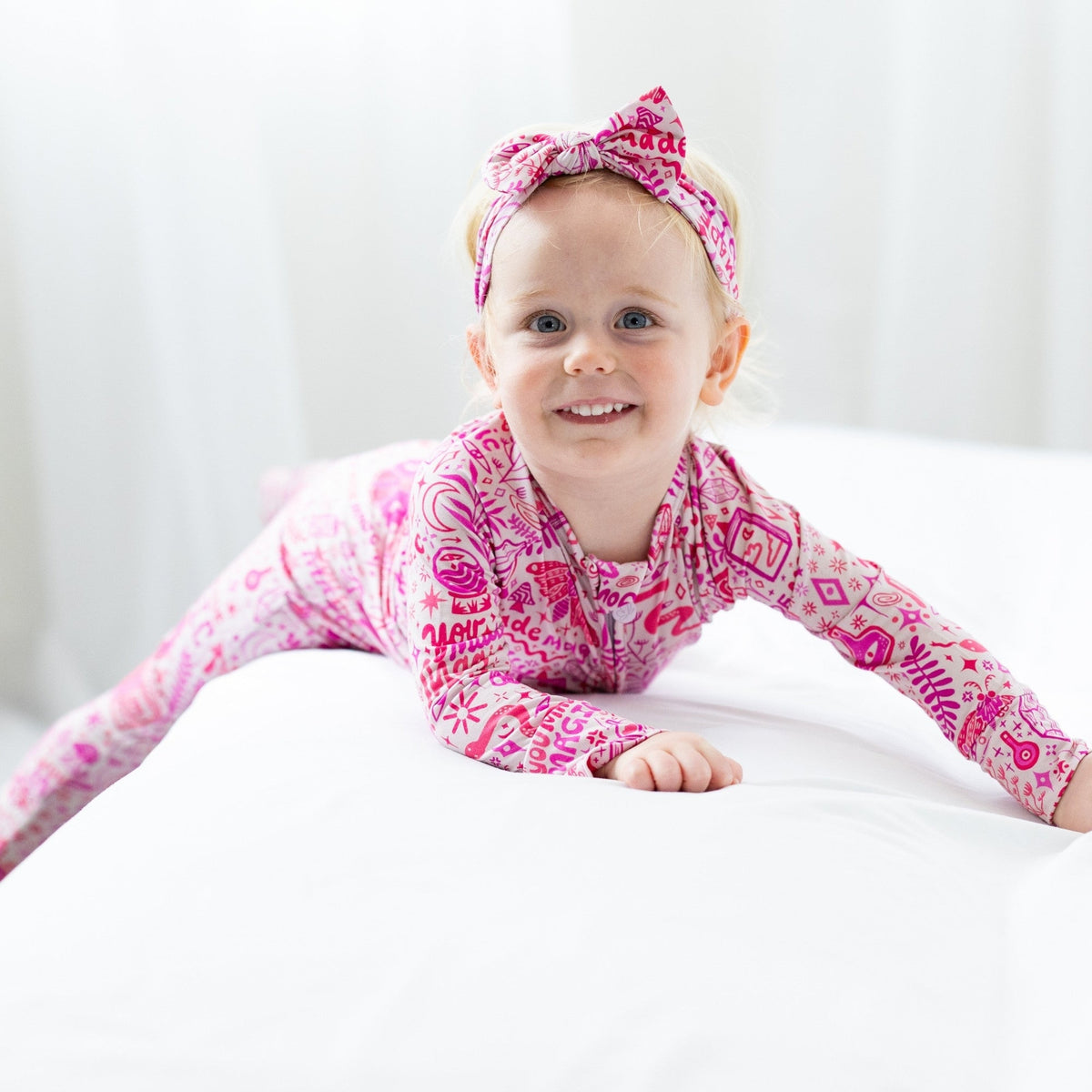Smiling toddler in a pink patterned romper and matching headband on a white bed.