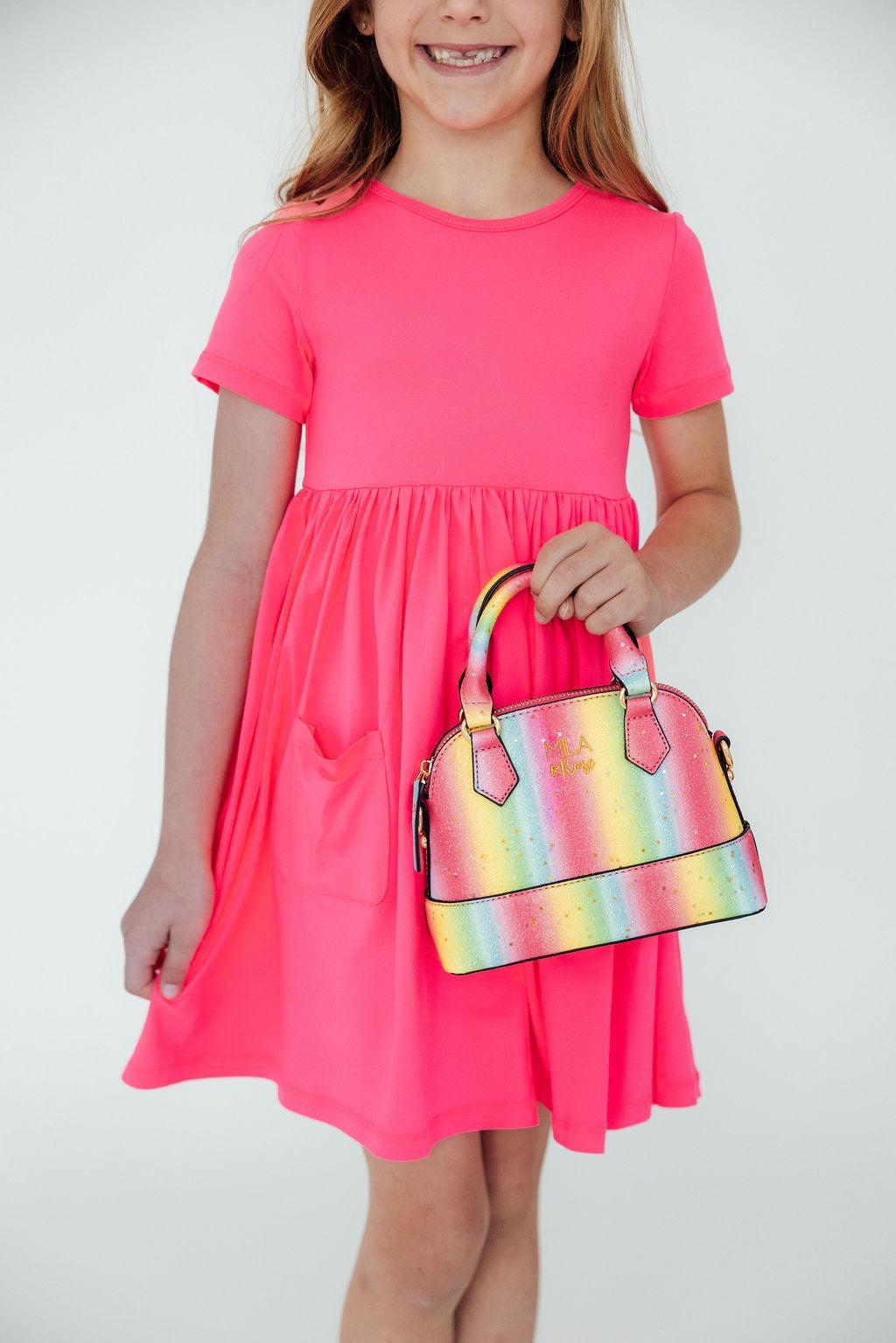 Girl in pink dress holding neon rainbow glitter crossbody purse. Sparkly fashion accessory for girls with handle and strap.