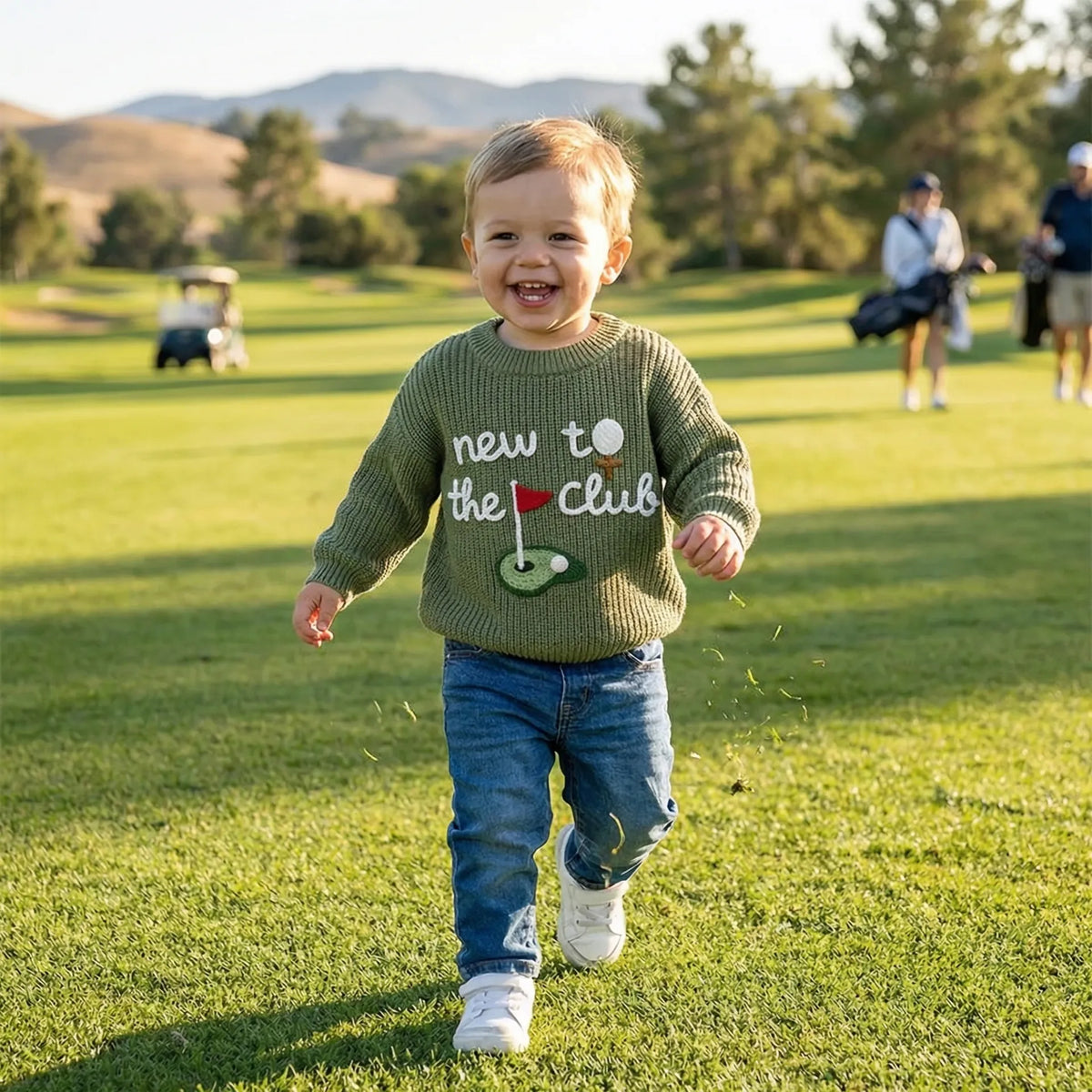 Toddler boy in olive green embroidered golf sweater running on grassy course with blurred mountains and carts in background