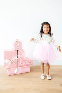 Girl wearing a festive long-sleeve tutu dress with a pink tulle skirt, standing next to a stack of pink wrapped gifts.