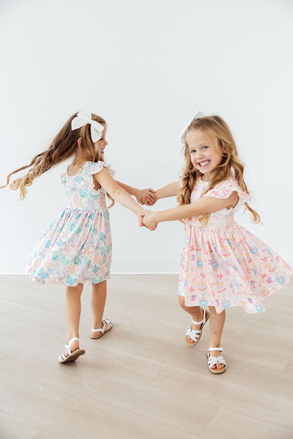 Two girls in pastel flutter-sleeve twirl dresses, holding hands and dancing.