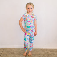 Om Baby light-blue floral two-piece short-sleeve pajama set on a smiling young girl, barefoot indoors.