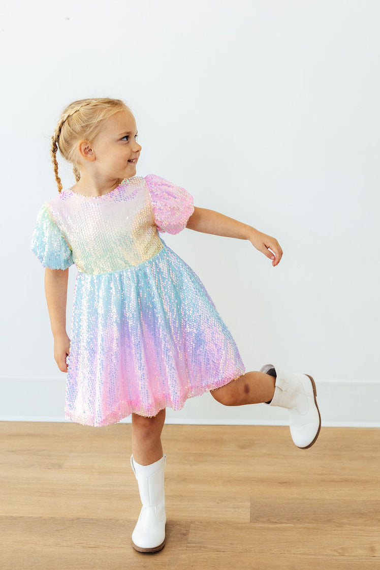 Toddler girl in rainbow ombre puff sleeve sequin dress and white boots, posing playfully for special occasions.