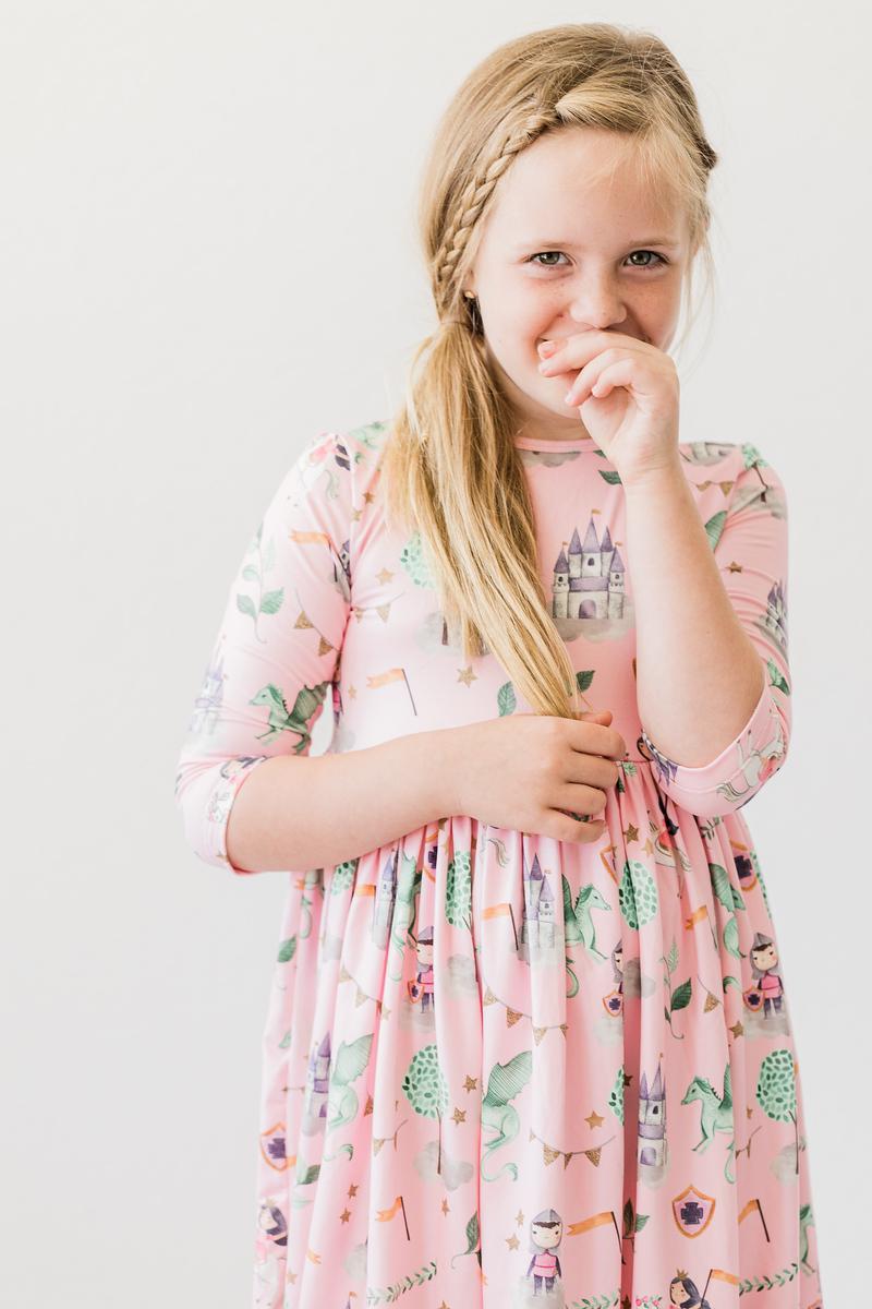 Smiling girl wearing the Once Upon a Time 3/4 Sleeve Pocket Twirl Dress by Mila & Rose, her cheerful expression brightening the whimsical dress adorned with fantastic designs.