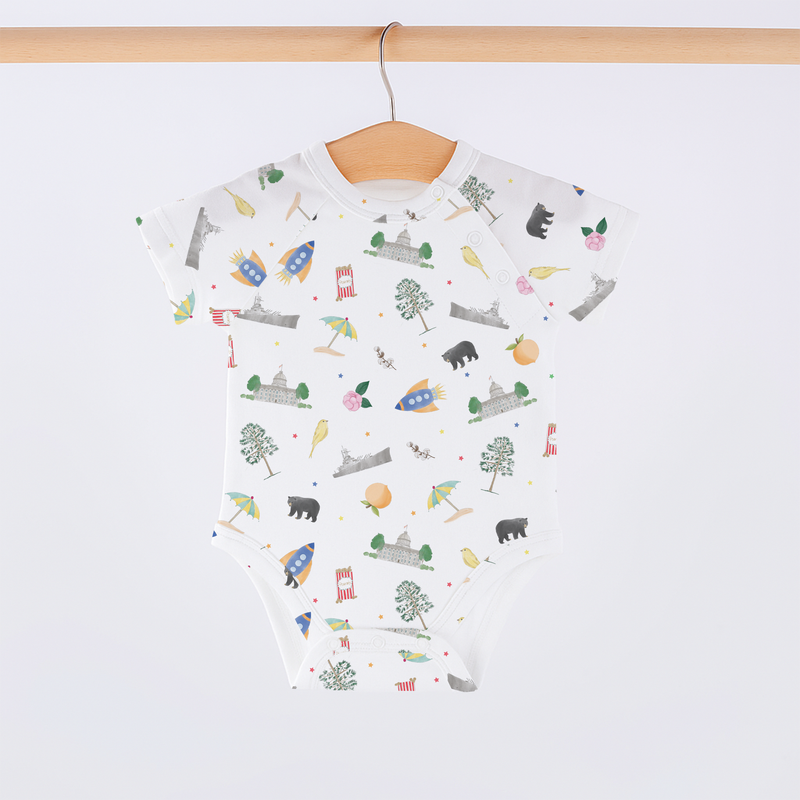 Alabama Kids Organic Cotton Onesie on hanger with lap-shoulder neck; white fabric features travel-themed prints.