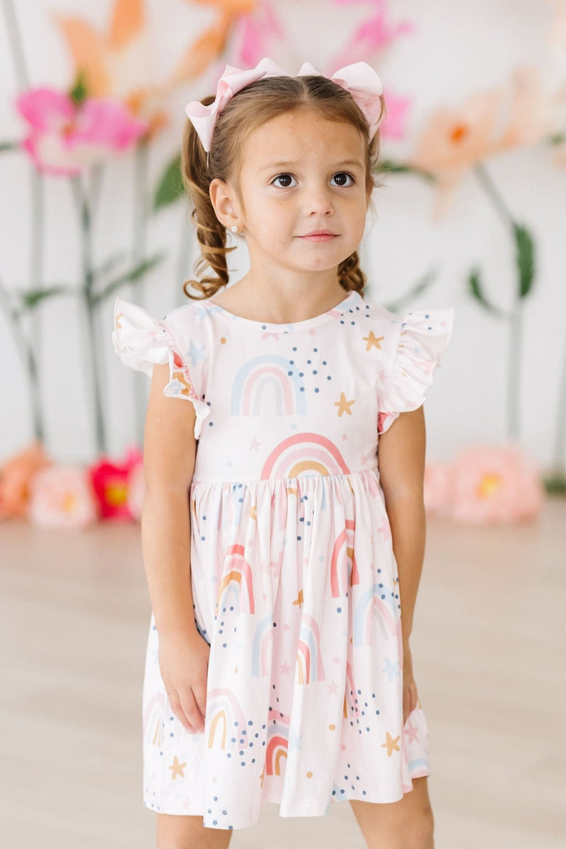 A young girl gazes thoughtfully while wearing the Over the Rainbow Flutter Sleeve Twirl Dress by Mila & Rose, perfectly capturing the dress's whimsical design and charming appeal.