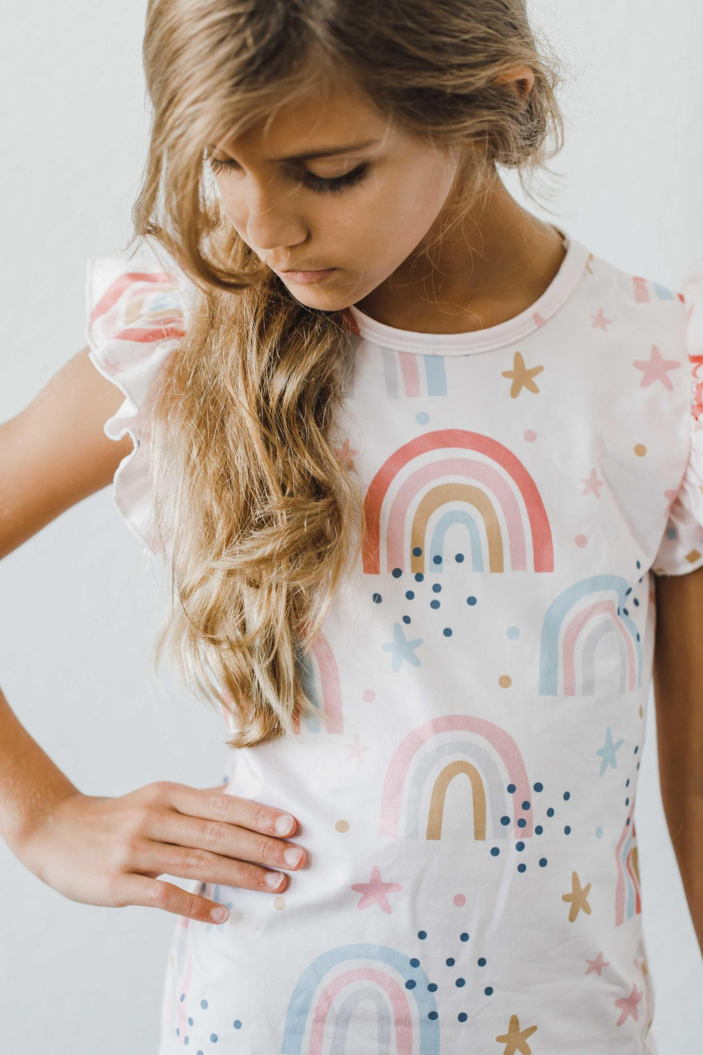 Over the Rainbow S/S Ruffle Tee in a playful design, showcasing a girl with long hair styled with waves posing thoughtfully, perfect for a stylish children's wardrobe.