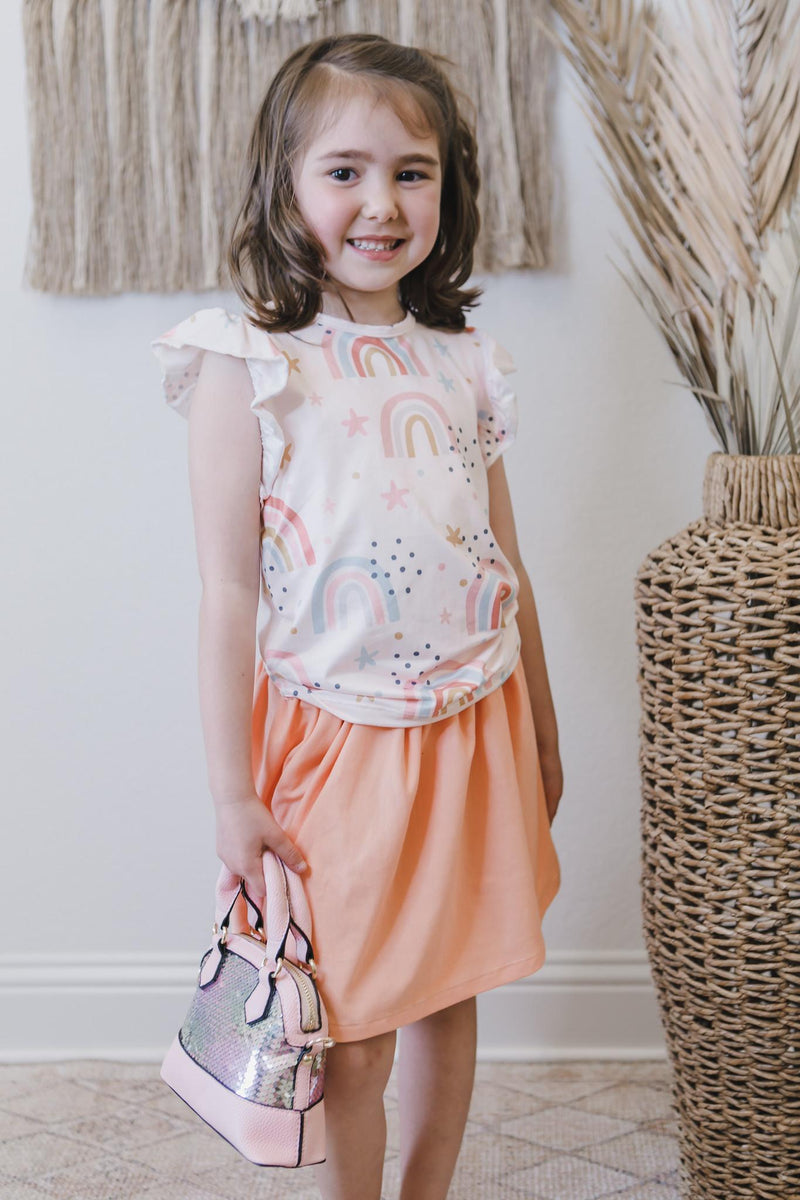 Over the Rainbow S/S Ruffle Tee worn by a smiling girl, paired with a cute peach skirt and a sparkly handbag, making every playdate a fashionable adventure.