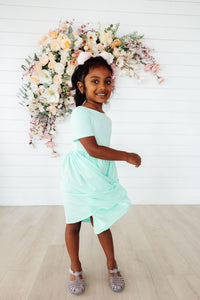Girl twirling in pastel green short-sleeve pocket twirl dress for girls, white bow, floral backdrop.