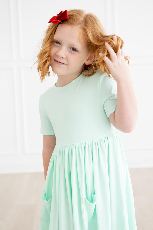 Pastel green short-sleeve pocket twirl dress for girls worn by a smiling red-haired girl with a red bow.