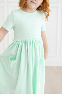 Girl wearing pastel green short-sleeve pocket twirl dress with a gathered waist and pockets.