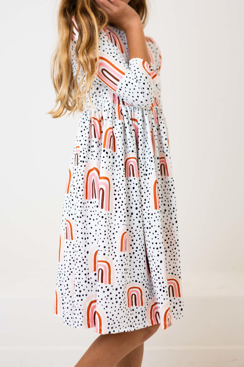 Young girl playfully poses in the Peach Rainbow 3/4 Sleeve Pocket Twirl Dress, showcasing its twirly design and eye-catching pattern of rainbows and polka dots.