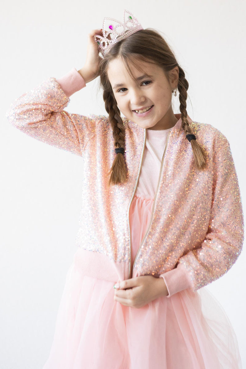 Young girl wearing a Peach Sequin Jacket, grinning and posing with a sparkling tiara, perfect for stylish playdates.