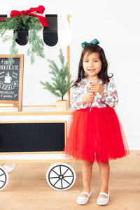 Little girl in candy cane print long sleeve top and red tulle tutu skirt holding a candy cane near a hot chocolate cart.