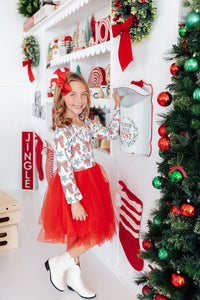 Girl in festive long sleeve dress with candy print top and red tutu skirt, posing near North Pole Post mailbox and Christmas tree.