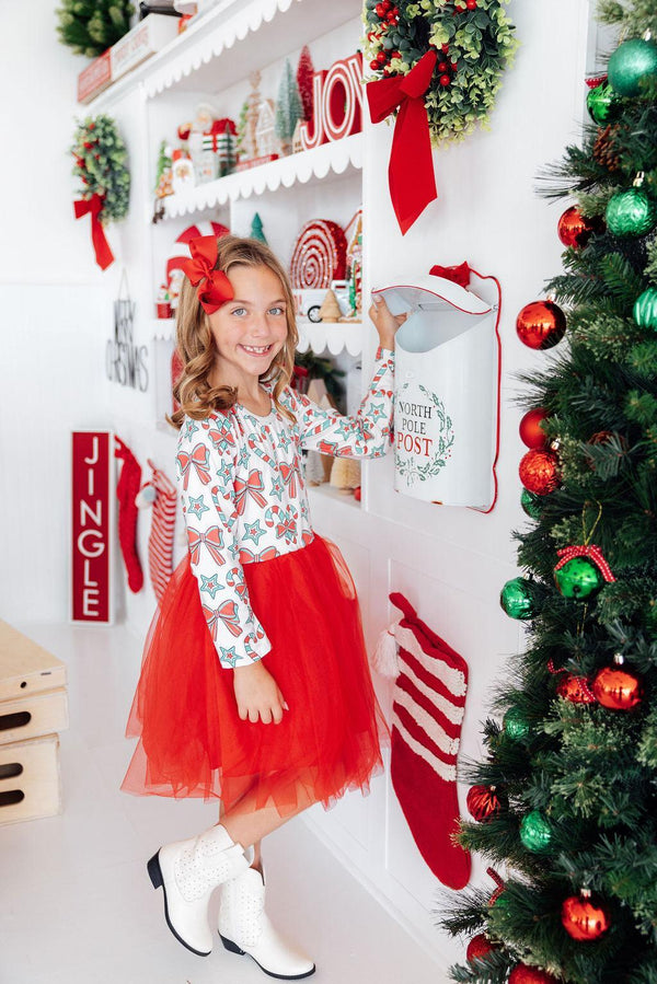 Girl in festive long sleeve dress with candy print top and red tutu skirt, posing near North Pole Post mailbox and Christmas tree.