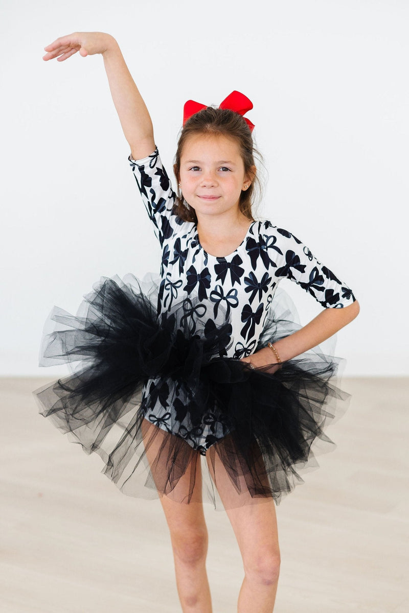 A young girl wearing the Perfectly Tied 3/4 Sleeve Tutu Leotard by Mila & Rose, showcasing a playful black and white bow design with a fluffy black tutu skirt. She is striking a pose with one arm raised and a red bow in her hair, perfect for dance classes or imaginative play.
