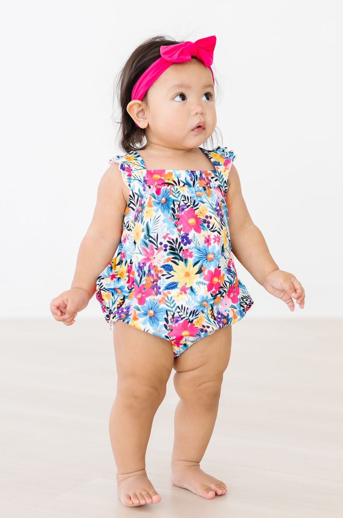 Toddler wearing Petal Paradise Ruffle Cross Back Romper in a bright floral print with a pink headband bow.