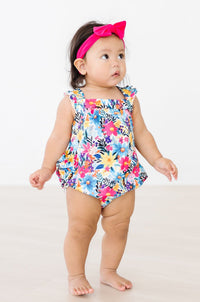 Toddler wearing Petal Paradise Ruffle Cross Back Romper in a bright floral print with a pink headband bow.