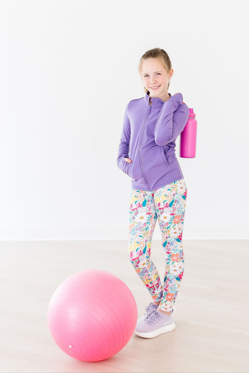 Girl smiling while holding a pink water bottle, dressed in the Playful Purple Full-Zip Ruffle Girl's Active Jacket next to an exercise ball.